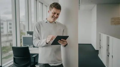 Justus is standing in an office environment in front of a column. He is holding a tablet in his hands. He is wearing a gray pullover and is smiling.