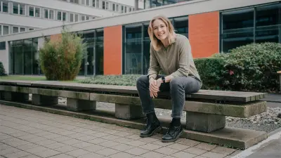 Katharina is sitting on a brown wooden bench in an outside area. Her arms are crossed as she is smiling at the camera. There is a building and plants in the background.