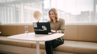 Katharina is sitting on a long bench at a table in front of her laptop. Next to her is a glass of water. In the background is a globe and a large window front.