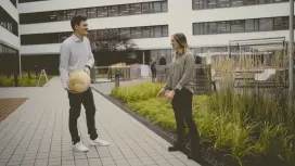 Justus and Katharina are standing opposite each other in an outside area. In the background, there is a building as well as green spaces and plants. They are throwing a small round globe between each other.