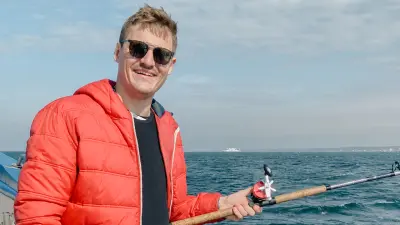 Justus is wearing a red, warm jacket and sunglasses. He is holding a fishing rod in his hands. In the background, one can see the sea.