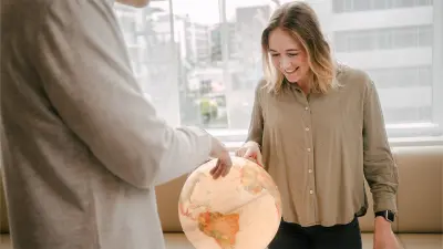 Katharina is looking at the globe in front of her with a smile. She is pointing a finger to a spot on the globe. In the foreground, a man’s upper body is depicted cropped and blurred. The man as well is pointing his right hand to the globe.