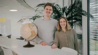 Justus und Katharina stehen nebeneinander an einem Tisch, sie blicken lächelnd in die Kamera. Links neben Justus steht ein Globus. Im Hintergrund ist eine Büroumgebung zu sehen, eine große grüne Pflanze ist hinter den beiden postiert.