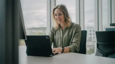 Katharina is sitting at a desk in front of a laptop, her hands placed on it. She is wearing a green blouse and is smiling.