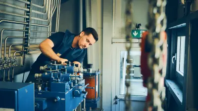 Technician working on machinery with pipes and control components inside an industrial facility.