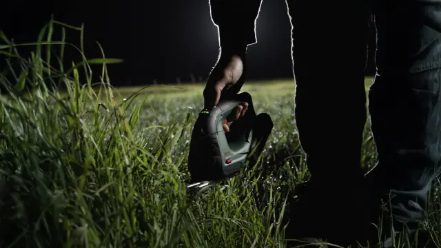 A man, slightly in the dark, bends down to cut longer blades of grass precisely with the Bosch AdvancedShear garden tool. The illuminated field in the background.
