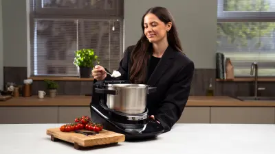 Frau steht hinter einem Cookit mit einem Löffel Eis und schaut diesen an. Im Vordergrund ist ein Brett mit Tomaten.