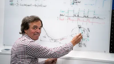 Tino in a checkered shirt smiles in front of a whiteboard covered in quantum physics equations and diagrams.
