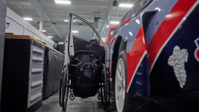 Außenaufnahme des Rennwagens auf der Fahrerseite, wo neben der geöffneten Fahrertür ein Rollstuhl steht.