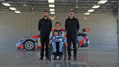 Robert Wickens im Rennfahreroutfit und mit Helm auf dem Schoß in seinem Rollstuhl, links und rechts von ihm stehen zwei Bosch Motorsport Mitarbeiter. Hinter ihnen der Rennwagen.