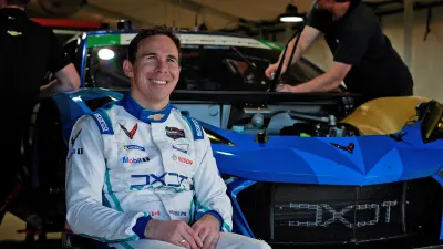 Robert Wickens im Rennfahreroutfit sitzt im Rollstuhl vor seiner Chevrolet Corvette Z06 TG3.R. Hinter ihm wird an dem Rennwagen gearbeitet.