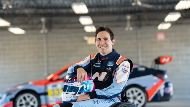 Robert Wickens smiles into the camera in his racing driver outfit and with his helmet on his lap, his racing car in the background.