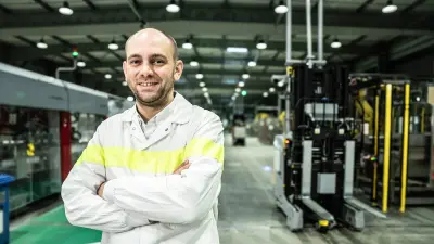 François Duquesne im weißen Kittel, er verschränkt die Armen und lächelt in die Kamera. Hinter ihm befindet sich eine große Produktionshalle mit Maschinen und einem Gabelstapler.