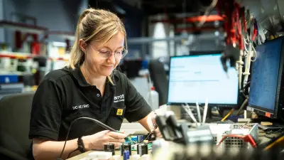 Eine Frau mit Schutzbrille lächelt, während sie eine Leiterplatte in der Hand hält und einen Lötkolben in der anderen Hand hält.