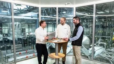 Drei Männer stehen und unterhalten sich in einem Besprechungsraum mit Blick auf die Fabrikhalle von Normandise Pet Food.
