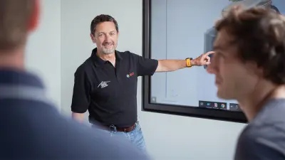 Reiner steht vor einem Monitor, zeigt darauf und bespricht sich mit seinen Kollegen.