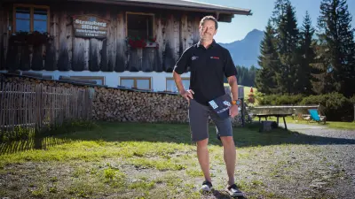 Reiner steht mit seinem Laptop in der Hand vor seiner Berghütte in den Allgäuer Alpen.