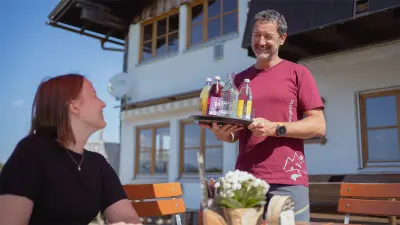 Reiner hält ein Tablett mit Getränken in der Hand und lächelt einen Gast an.