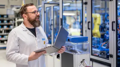 Person in lab coat working on a laptop in automated machinery surroundings.