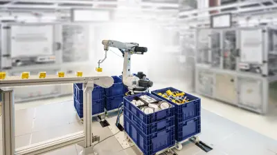 Zu sehen ist ein schwenkbarer Roboterarm von Bosch Rexroth in einem Fabrikumfeld. Darüber ist eine Kamera montiert. Der Roboterarm greift mit einem am Greifarm montierten Saugnapf eine kleine Box aus einer sortenreinen Kiste und legt ihn auf das Förderband.