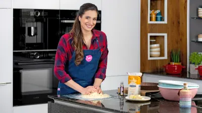 ie Food-YouTuberin Sally Özcan steht in ihrer Küche hinter einer Arbeitsfläche, knetet einen Teig und lächelt in die Kamera.