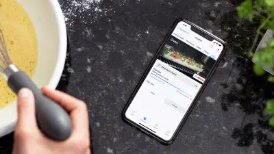 Ein Smartphone, das durch die Vernetzung mit der Home-Connect-App den Backfortschritt einer Tarte anzeigt, liegt auf einer Arbeitsfläche neben einer Rührschüssel, in der die Food-YouTuberin Sally Özcan eine Creme zubereitet.