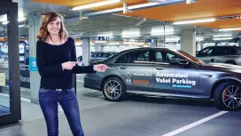 Im Parkhaus des Mercedes-Benz Museums in der Stadt Stuttgart steht Sabine Sayler und zeigt mit beiden Händen auf einen Mercedes mit der Außenaufschrift Automated Valet Parking.