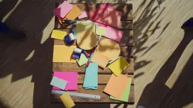 There are many colourful pieces of paper in the middle of a table. Several people are standing around the table, visible from above. They are grabbing the pieces of paper.