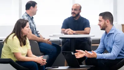 Das Testimonial Guilherme sitzt gemeinsam mit einer Frau an einem Tisch. Sie befinden sich in einem Gespräch. Vor Guilherme steht ein Laptop. Im Hintergrund sind zwei weitere Männer an einem Tisch zu sehen, die sich ebenfalls in einem Gespräch befinden.