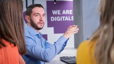 Das Testimonial Guilherme sitzt an einem Tisch, vor ihm befindet sich ein Laptop. Im Vordergrund sind zwei Frauen von hinten zu sehen, mit denen er im Gespräch ist. Er gestikuliert mit den Händen. Im Hintergrund erkennt man eine Wand, auf der sich der Schriftzug „We are Digitals” befindet.