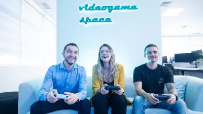 Das Testimonial Guilherme sitzt neben einer Frau und einem weiteren Mann auf einem Sofa. Sie alle lächeln und haben einen Spielkonsolen-Controller in der Hand. Im Hintergrund ist der Schriftzug „Videogama Space” zu lesen.