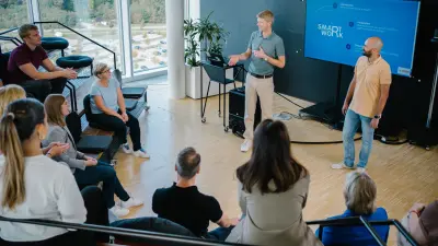 Das Testimonial Joachim und das Testimonial Stefan befinden sich in einer Workshop-Umgebung. Hinter ihnen ist ein großer Bildschirm zu sehen, auf dem „Smart Work” zu 20 lesen ist. Vor ihnen befindet sich eine Gruppe aus rund 9 Menschen, die in einem Halbkreis um sie herum sitzen und ihre Blicke auf die beiden richten.