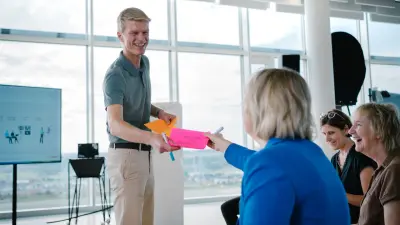 Das Testimonial Joachim ist in einer Workshop-Umgebung zu sehen. Er steht vor mehreren Stühlen, auf denen drei Teilnehmerinnen sitzen. Er gibt einer der Teilnehmerinnen lächelnd bunte Karten in die Hand.