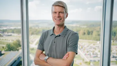 Das Testimonial Joachim befindet sich vor einem Fenster in einem oberen Stockwerk einer Büroumgebung. Er ist bis zum Oberkörper zu sehen, hat die Arme verschränkt und lächelt in die Kamera.