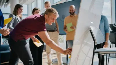 Mehrere Frauen und Männer stehen um ein Whiteboard herum. Ein Mann pinnt lächelnd einen Zettel daran.