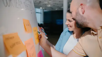 Ein Mann und eine Frau sind bis zum Oberkörper von der Seite zu sehen. Sie lächeln und befinden sich vor einem Whiteboard. Beide haben Stifte in der Hand und schreiben etwas auf orange Zettel, die sich auf dem Whiteboard befinden.