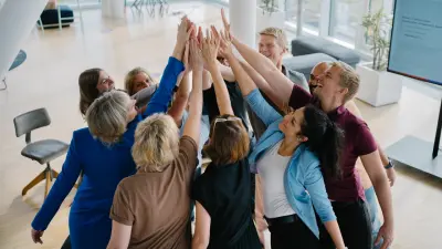 Mehrere Männer und Frauen stehen in einem Kreis zusammen. Sie alle heben eine Hand in die Mitte zu einem High-Five. Sie befinden sich in einer Büroumgebung mit einer Workshop-Fläche im Hintergrund.