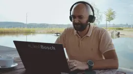 Das Testimonial Stefan sitzt in einer Außenumgebung an einem Holztisch. Im Hintergrund ist ein größerer Teich und eine Grünfläche zu sehen. Er hat einen Laptop vor sich, den er aufklappt. Daneben befinden sich Kopfhörer und eine Kaffeetasse auf dem Tisch.