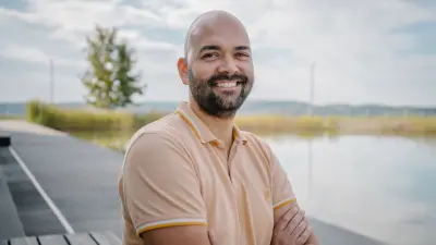 Das Testimonial Stefan ist bis zum Oberkörper zu sehen. Er lächelt in die Kamera und hat die Arme verschränkt. Er befindet sich in einer Außenumgebung, im Hintergrund ist ein größerer Teich und Grünfläche zu sehen.