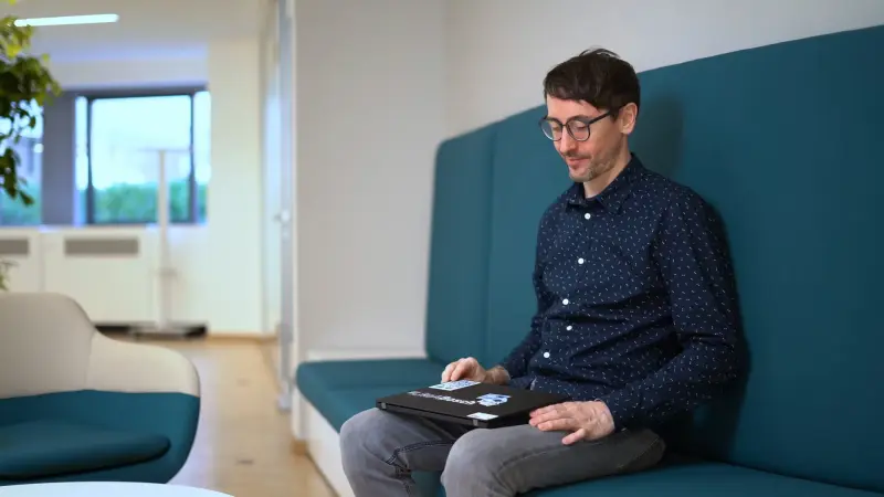 Enrico sits on a petrol-coloured sofa and opens and closes his laptop, which he has on his lap.