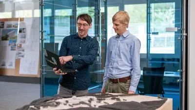 Enrico and his colleague stand in front of a model and look at the laptop Enrico holds.
