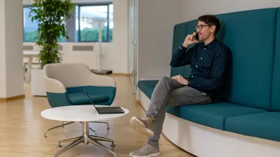 Enrico sits on a petrol-coloured sofa and talks on the phone; his laptop is on a table in front of him.