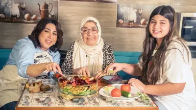 Bassent Helmy sits around a square table on the left, her mother in the middle and her daughter on the right. The family is preparing a salad together and chopping different types of vegetables, which come together in a large glass bowl.