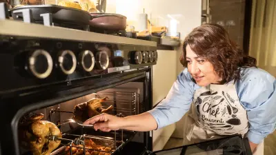 The open F290 Ranger Cooker can be seen on the left. The oven door is half open, two roast chickens on skewers can be seen in the oven pipe. A woman on the right is kneeling at the height of the oven and testing with a fork whether the meat is cooked.