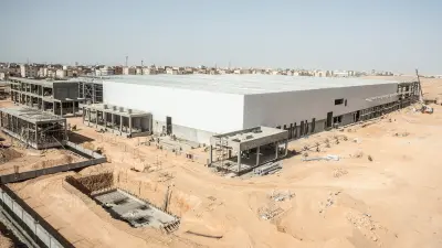 A bird's eye view of the construction site of the future BSH stove plant in Ramandan City. The light is bright and with a blue sky and yellow sandy soil. The front part is already clad with white outer walls, on the left of the picture you can still see open parts of the building shell.