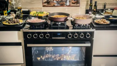 The black glazed FS90 Range Cooker forms the center of the picture. In the oven itself two grilled chickens are roasted on skewers, with vegetables cooking underneath. Four more dishes are being prepared on the gas hobs. To the left and right of the stove, the kitchen worktop is filled with other ingredients for the dishes being prepared.