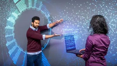 Two people stand in front of a screen projection of many small spheres. One person has a laptop in her hand, from which a kind of data cloud is wafting.