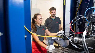Bosch engineers Tsvetelina Yordanova (left) and Peter Stritt (right) standing in front of a factory test stand and testing the laser welding prototype at the Bosch Research site in Renningen. A system with numerous cables can be seen.