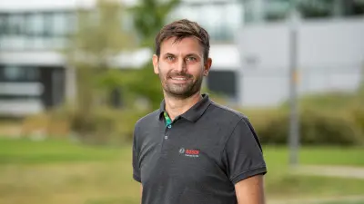 Bosch Research mechanical engineer Peter Stritt in a Bosch polo shirt can be seen in the foreground. The Bosch Research Renningen site is visible, blurred, in the background.