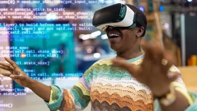 : A young man is wearing virtual reality goggles and is excitedly discovering a mysterious world. He has his arms stretched out wide. A digital Keras code of numbers and letters buzzes around him, intended to illustrate the artificial intelligence behind it.